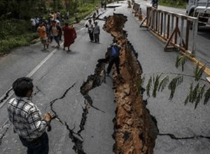 A large crack runs through the middle of a paved road, caused by an earthquake. People gather around the damaged area, inspecting the deep fissure and the surrounding destruction.